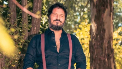 Hombre de cabello oscuro y barba, vestido con camisa negra y tirantes granates, posando al aire libre entre árboles.