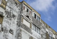 Fachada deteriorada de un bloque de viviendas con ventanas, celosías y una abertura irregular en el revestimiento exterior.