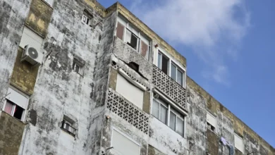 Fachada deteriorada de un bloque de viviendas con ventanas, celosías y una abertura irregular en el revestimiento exterior.