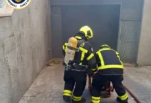 Dos bomberos de espaldas, con equipos de protección y respiración, junto a la entrada de un garaje donde se aprecia humo.