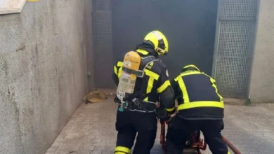 Dos bomberos de espaldas, con equipos de protección y respiración, junto a la entrada de un garaje donde se aprecia humo.