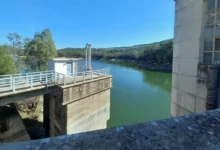 Vista de una infraestructura de hormigón con pasarelas y barandillas metálicas junto al agua verde de un embalse, rodeado de vegetación.