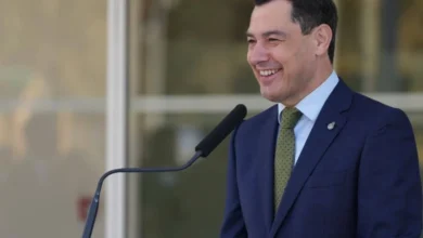 Juanma Moreno sonríe junto a un micrófono durante una intervención institucional, vestido con traje azul oscuro, camisa clara y corbata verde.