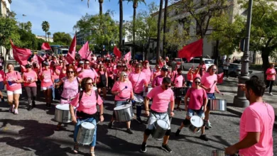 Participantes vestidos de rosa durante una marcha solidaria por una calle arbolada, con banderas rosas y músicos con tambores.