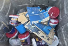 Varios bidones y envases de pintura, tapas rojas, cartones azules y bolsas acumulados en el interior de un contenedor de basura.