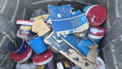 Varios bidones y envases de pintura, tapas rojas, cartones azules y bolsas acumulados en el interior de un contenedor de basura.