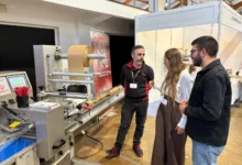Tres personas conversan junto a una máquina industrial con un gran rollo de material de color marrón en un recinto de exposiciones.