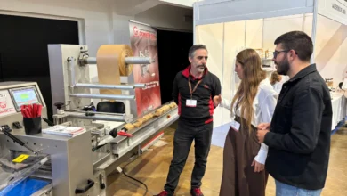 Tres personas conversan junto a una máquina industrial con un gran rollo de material de color marrón en un recinto de exposiciones.