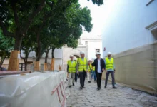 Personas con cascos y chalecos reflectantes caminan por una zona de obras con pavimento de piedra, árboles protegidos y muros blancos.