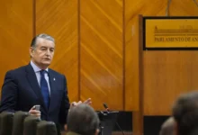 Un hombre con traje oscuro y corbata azul interviene ante varios asistentes en una sala con el rótulo Parlamento de Andalucía.