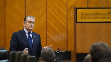 Un hombre con traje oscuro y corbata azul interviene ante varios asistentes en una sala con el rótulo Parlamento de Andalucía.