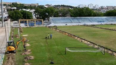 Vista general de un campo de fútbol con zonas de tierra abiertas junto al césped, una miniexcavadora amarilla, dos personas sobre el terreno y gradas azules al fondo.