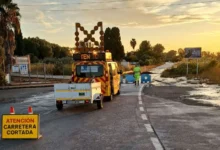 Vehículo amarillo de conservación de carreteras con remolque, señal de carretera cortada y un operario junto a una calzada mojada.