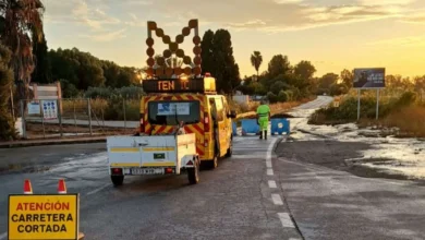 Vehículo amarillo de conservación de carreteras con remolque, señal de carretera cortada y un operario junto a una calzada mojada.