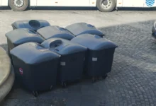 Grupo de contenedores de basura oscuros con tapa cerrada, situados sobre un pavimento de adoquines junto a un autobús y una furgoneta.