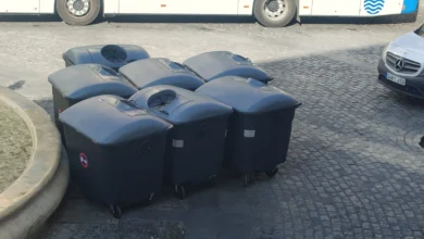 Grupo de contenedores de basura oscuros con tapa cerrada, situados sobre un pavimento de adoquines junto a un autobús y una furgoneta.