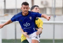 Dos futbolistas disputan un balón, con un jugador de camiseta azul y pantalón blanco por delante de otro vestido de amarillo y verde.