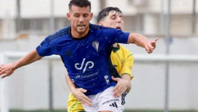 Dos futbolistas disputan un balón, con un jugador de camiseta azul y pantalón blanco por delante de otro vestido de amarillo y verde.