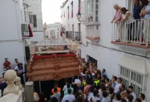 Paso procesional de madera tallada con una figura yacente y flores rojas rodeado de público en una calle estrecha de fachadas blancas.