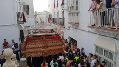 Paso procesional de madera tallada con una figura yacente y flores rojas rodeado de público en una calle estrecha de fachadas blancas.