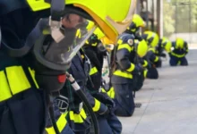 Bomberos equipados con cascos amarillos, máscaras respiratorias y trajes oscuros con bandas reflectantes, arrodillados en una fila.