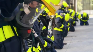 Bomberos equipados con cascos amarillos, máscaras respiratorias y trajes oscuros con bandas reflectantes, arrodillados en una fila.