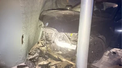 Frontal de un coche oscuro con daños graves tras impactar contra una estructura y una columna.