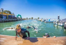 Varios nadadores con gorros y gafas de natación llegan a un pantalán de madera en un puerto deportivo con embarcaciones amarradas.