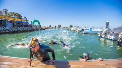 Varios nadadores con gorros y gafas de natación llegan a un pantalán de madera en un puerto deportivo con embarcaciones amarradas.