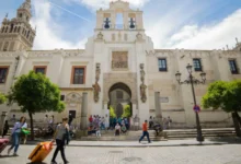 Fachada de un edificio histórico de arquitectura monumental con torres, campanario y un acceso central, ante el que caminan varias personas con equipaje.