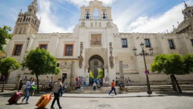 Fachada de un edificio histórico de arquitectura monumental con torres, campanario y un acceso central, ante el que caminan varias personas con equipaje.