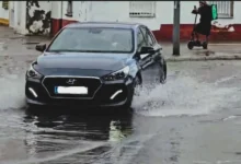 Turismo oscuro circulando por una calle con agua acumulada y levantando salpicaduras a ambos lados del vehículo.