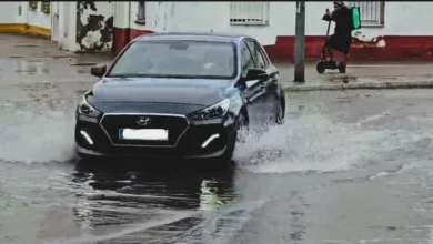 Turismo oscuro circulando por una calle con agua acumulada y levantando salpicaduras a ambos lados del vehículo.