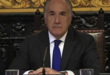 Hombre de traje oscuro, camisa blanca y corbata azul, sentado ante un micrófono con las manos apoyadas sobre una mesa.
