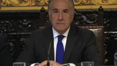 Hombre de traje oscuro, camisa blanca y corbata azul, sentado ante un micrófono con las manos apoyadas sobre una mesa.
