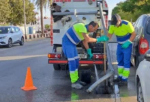 Dos operarios con ropa de alta visibilidad trabajan junto a una alcantarilla abierta, con una manguera y un camión de saneamiento estacionado detrás.