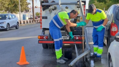 Dos operarios con ropa de alta visibilidad trabajan junto a una alcantarilla abierta, con una manguera y un camión de saneamiento estacionado detrás.
