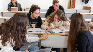Cinco estudiantes sentados alrededor de una mesa de aula, consultando libros y apuntes mientras estudian en grupo.
