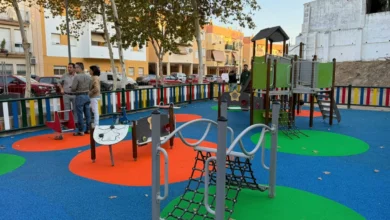 Parque infantil con suelo de caucho azul, zonas circulares verdes y naranjas, estructuras de juego y valla multicolor.