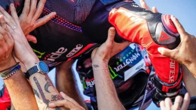 Primer plano de un piloto de motociclismo con mono de competición negro y rojo, rodeado de manos que lo felicitan.