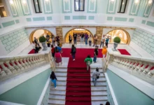 Vista elevada de una escalera de mármol con alfombra roja y varias personas en un amplio interior decorado en tonos verde claro y blanco.