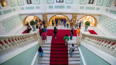 Vista elevada de una escalera de mármol con alfombra roja y varias personas en un amplio interior decorado en tonos verde claro y blanco.