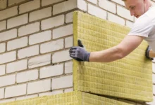 Trabajador con guantes oscuros colocando un panel amarillo de aislamiento térmico sobre una pared de bloques claros.