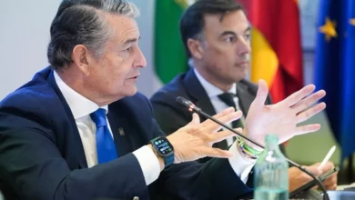 Dos hombres vestidos con traje participan en una reunión, con uno de ellos hablando y gesticulando ante un micrófono.