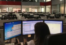 Persona vista de espaldas ante tres monitores en una amplia sala de trabajo con numerosas pantallas y puestos informáticos.