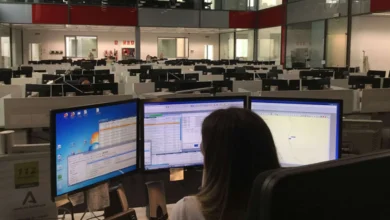 Persona vista de espaldas ante tres monitores en una amplia sala de trabajo con numerosas pantallas y puestos informáticos.