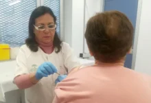 Profesional sanitaria con guantes administra una vacuna en el brazo de una persona sentada de espaldas en una consulta.