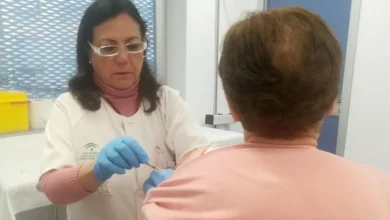Profesional sanitaria con guantes administra una vacuna en el brazo de una persona sentada de espaldas en una consulta.