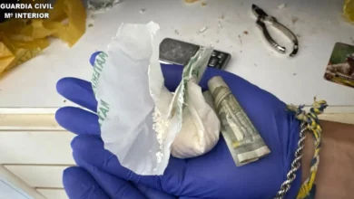 Manos con guantes azules sostienen envoltorios con una sustancia blanca en polvo y un billete enrollado.