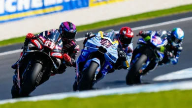Tres pilotos de motociclismo compiten en una curva, con una moto negra y roja delante de otras dos motos azules.